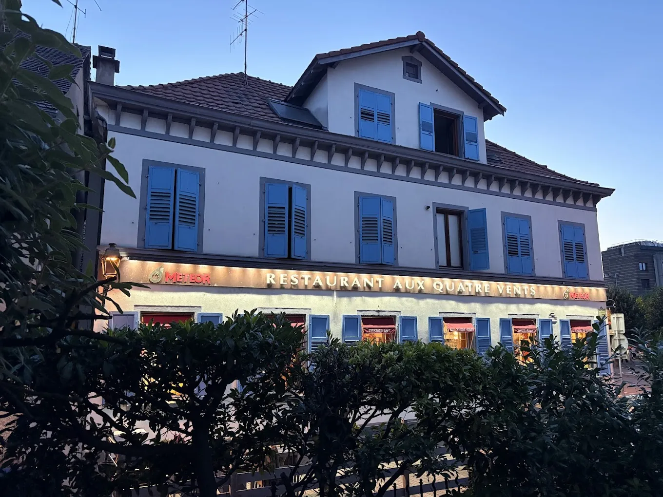 La façade blanche aux volets bleus du restaurant Aux Quatre Vents, enseigne allumée à la tombée de la nuit
